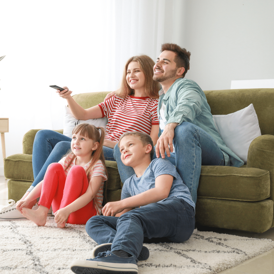 Untitled design (16) A family of four sits together in a cozy Sunshine Coast living room. The parents are on a green couch; the mother holds a remote control, while the father sits to her right. Two children, a girl and a boy, sit on the floor in front of them, all smiling and looking at a TV.