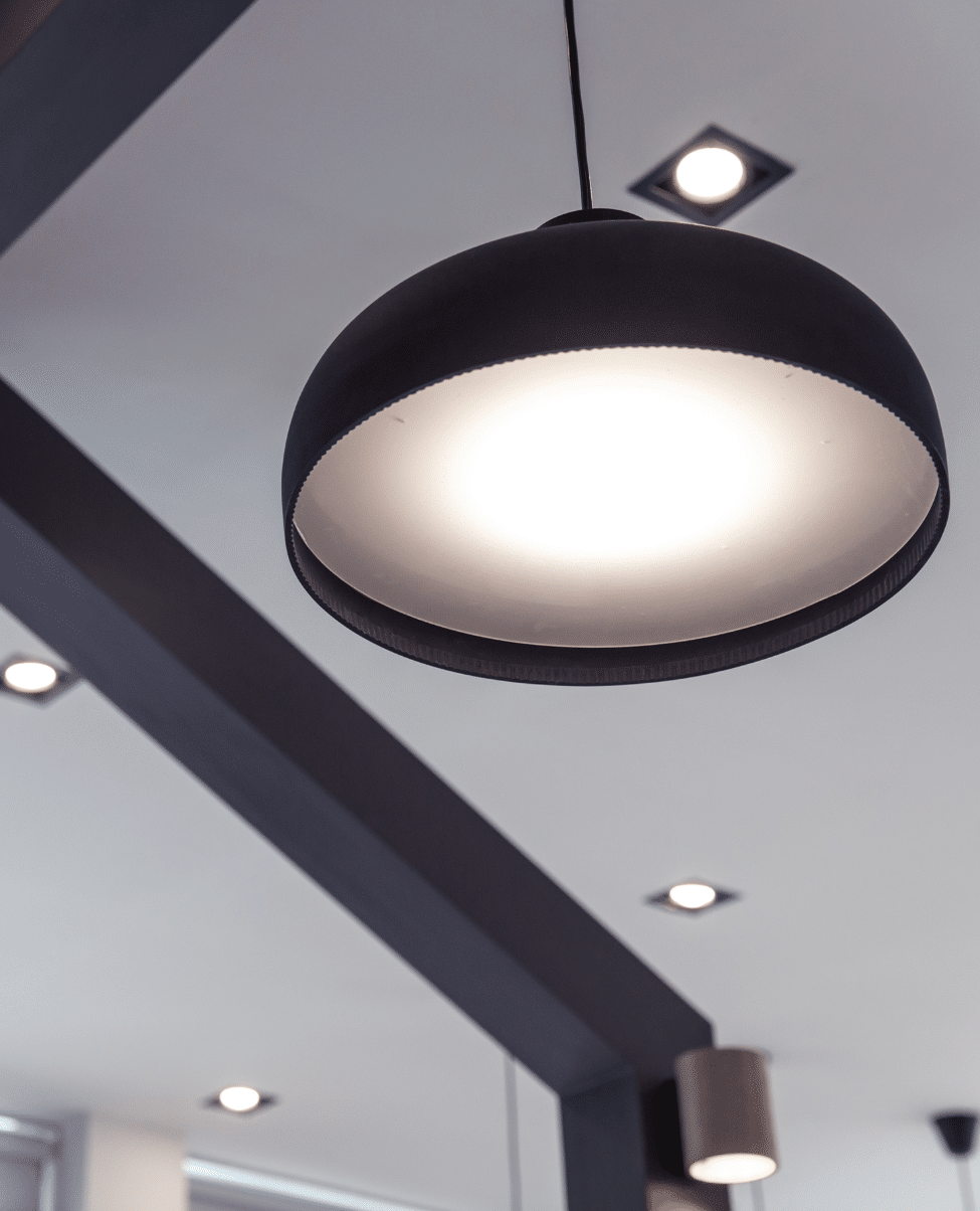 Untitled design (2) A modern hanging pendant light fixture is illuminated, casting a soft glow reminiscent of the Sunshine Coast. The light has a black exterior and a white interior, suspended from the ceiling by a cord. Several recessed ceiling lights are also visible in the background.