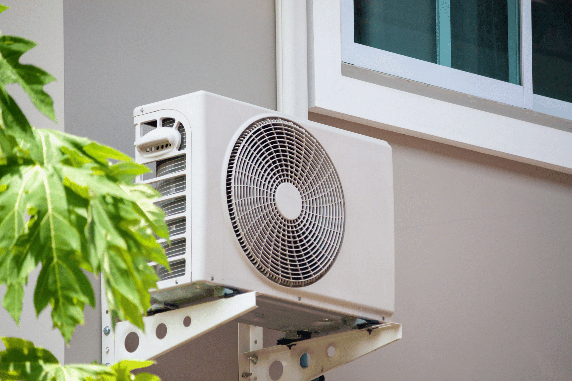 Untitled design - 2024-07-30T203152.025 A white outdoor air conditioning unit is mounted on the exterior wall of a building near a window, with green leaves partially obscuring the view. Situated on the Sunshine Coast, this serene setup blends modern convenience with natural beauty.