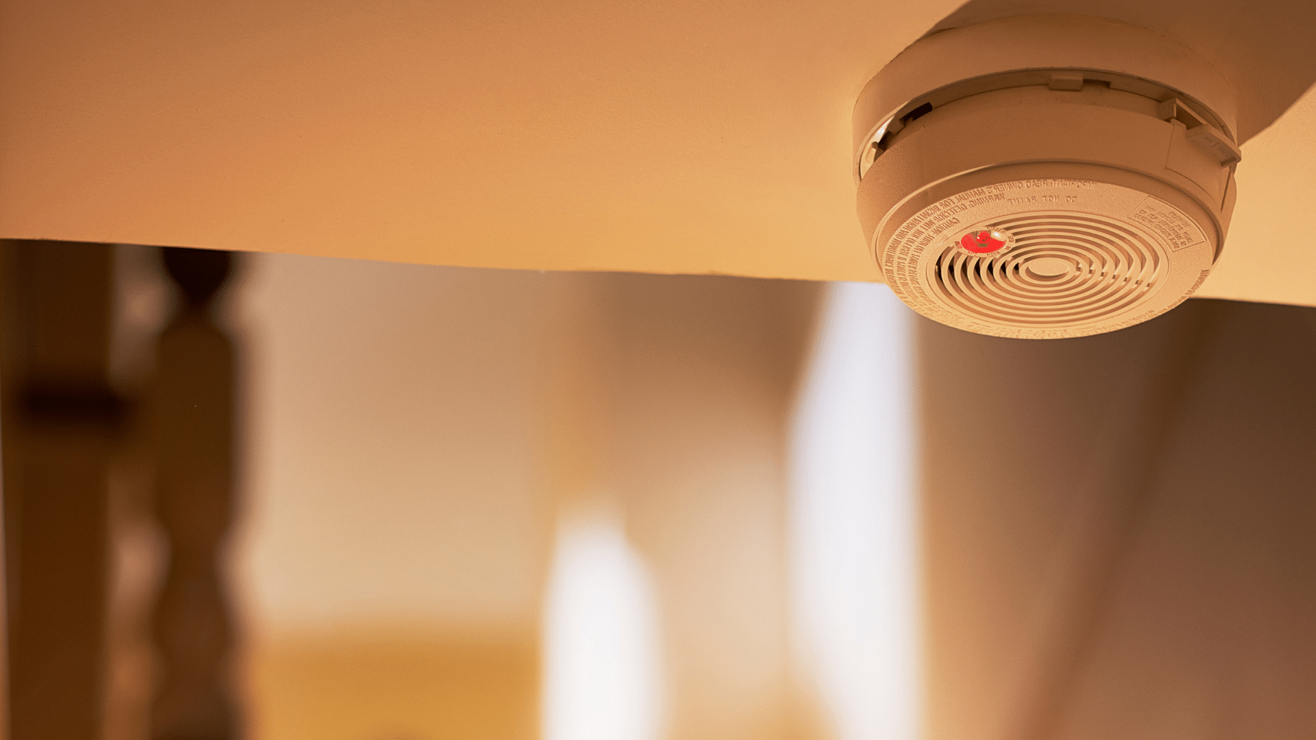 A close-up of a smoke detector mounted on a ceiling, featuring a glowing red indicator light, set against a blurred background with warm lighting reminiscent of the Sunshine Coast and a partially visible wooden railing on the left.