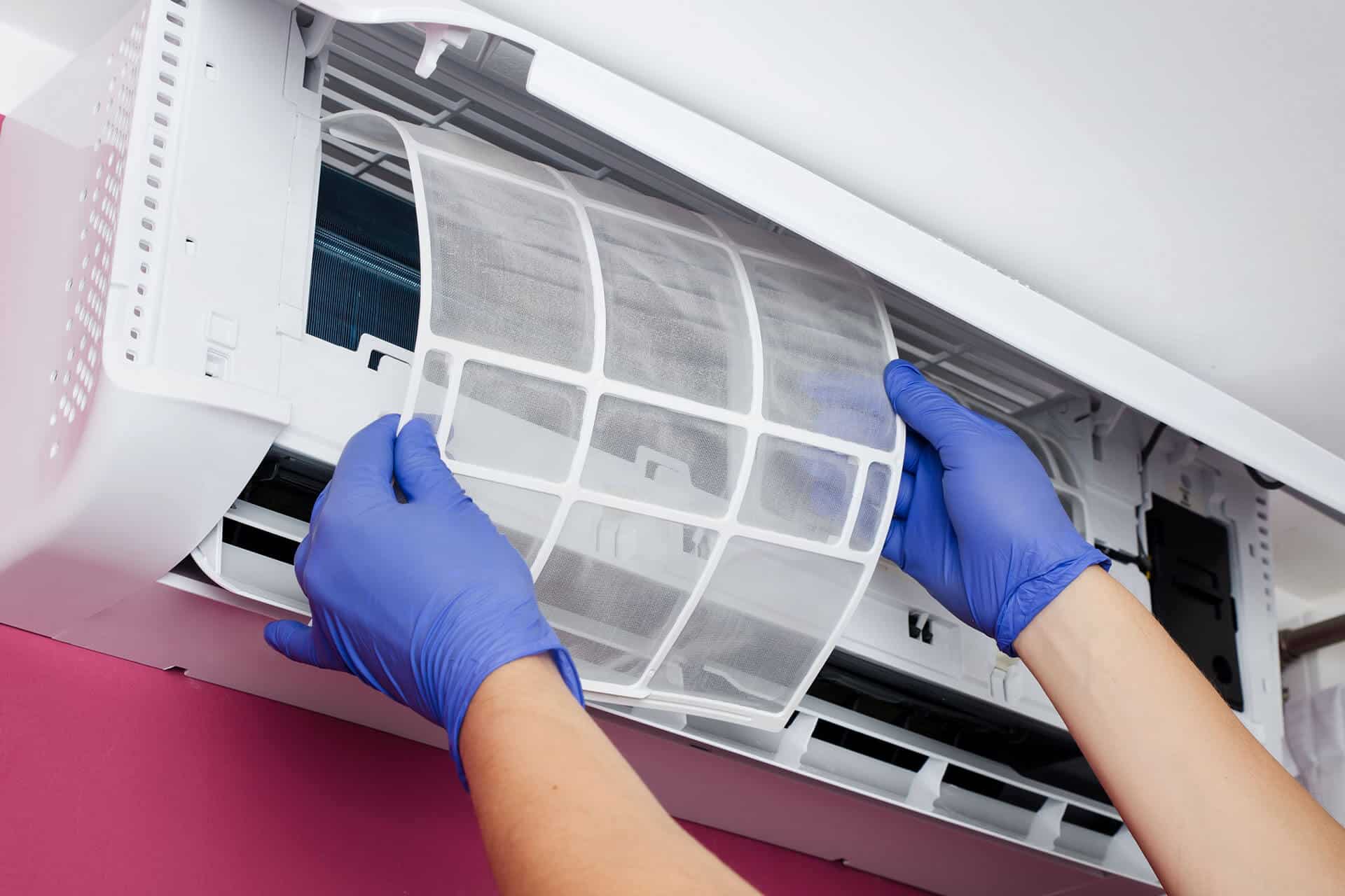 A person wearing blue gloves is removing a mesh air filter from a wall-mounted air conditioning unit. The unit is open, revealing its internal components against the backdrop of a pink-painted wall, reminiscent of the vibrant hues often seen on the Sunshine Coast.