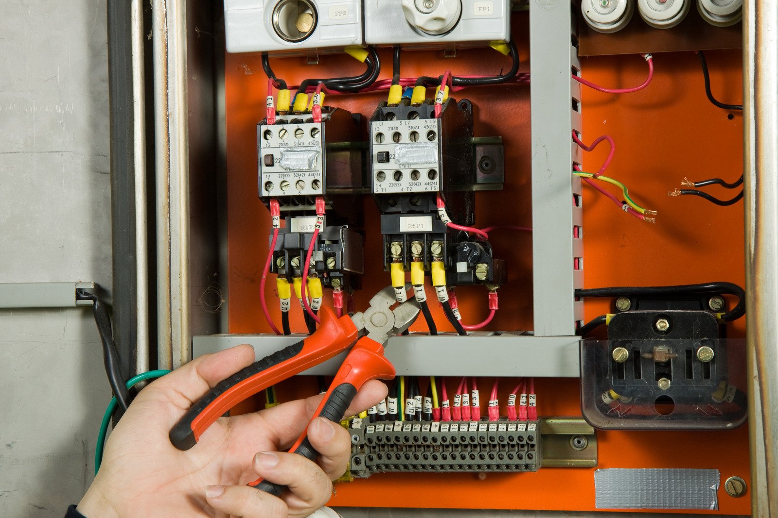 A hand holding red-handled wire cutters is positioned in front of an open electrical control panel with exposed wires, switches, and circuit components. A hand holding red-handled wire cutters is positioned in front of an open electrical control panel with exposed wires, switches, and circuit components.