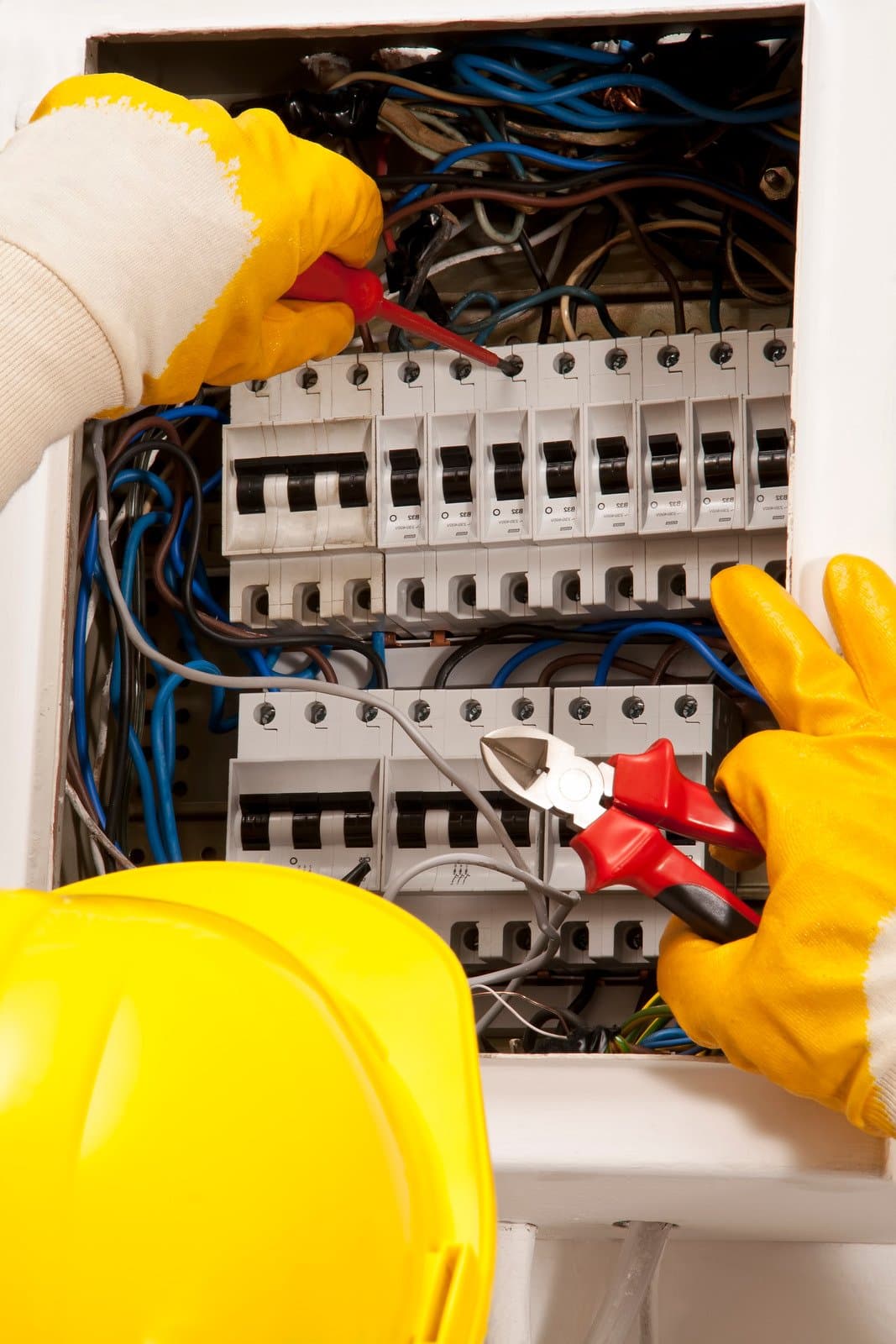 An electrician wearing yellow gloves and a yellow hard hat works on an open electrical panel, adjusting wires and circuit breakers with pliers and a screwdriver. An electrician wearing yellow gloves and a yellow hard hat works on an open electrical panel, adjusting wires and circuit breakers with pliers and a screwdriver.