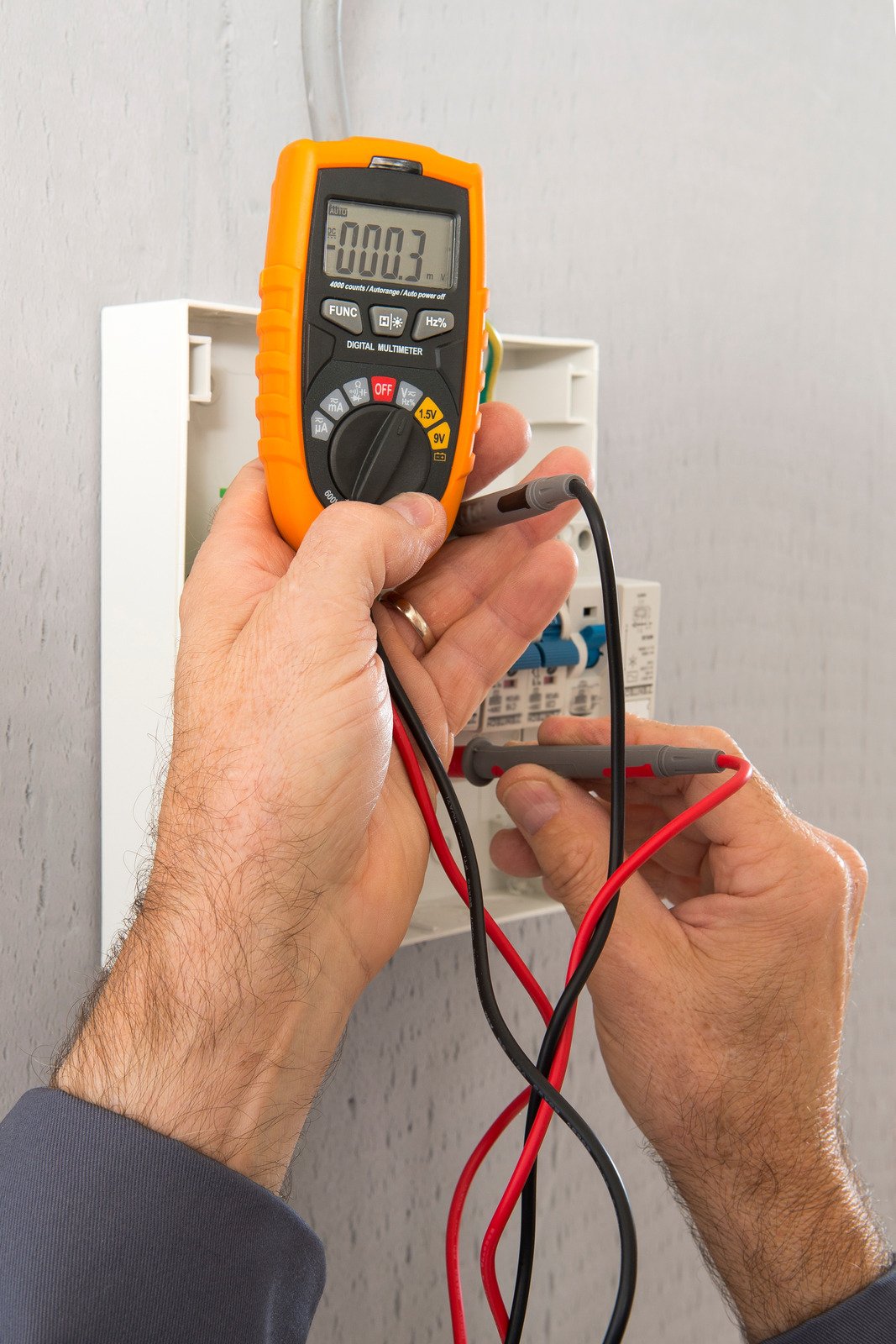 A person uses a digital multimeter to measure voltage or current inside an electrical panel, holding the meter probes to terminals while the display reads 0.03. A person uses a digital multimeter to measure voltage or current inside an electrical panel, holding the meter probes to terminals while the display reads 0.03.