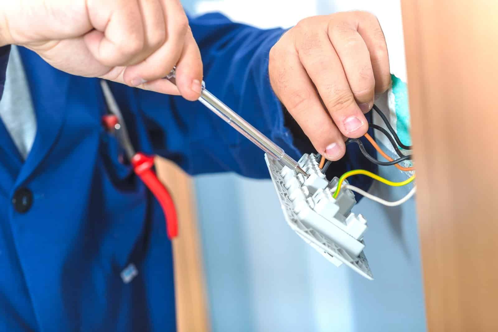 A person wearing a blue jacket uses a screwdriver to install or repair electrical wiring in a wall outlet. Several colored wires are visible, and pliers hang from their pocket. A person wearing a blue jacket uses a screwdriver to install or repair electrical wiring in a wall outlet. Several colored wires are visible, and pliers hang from their pocket.