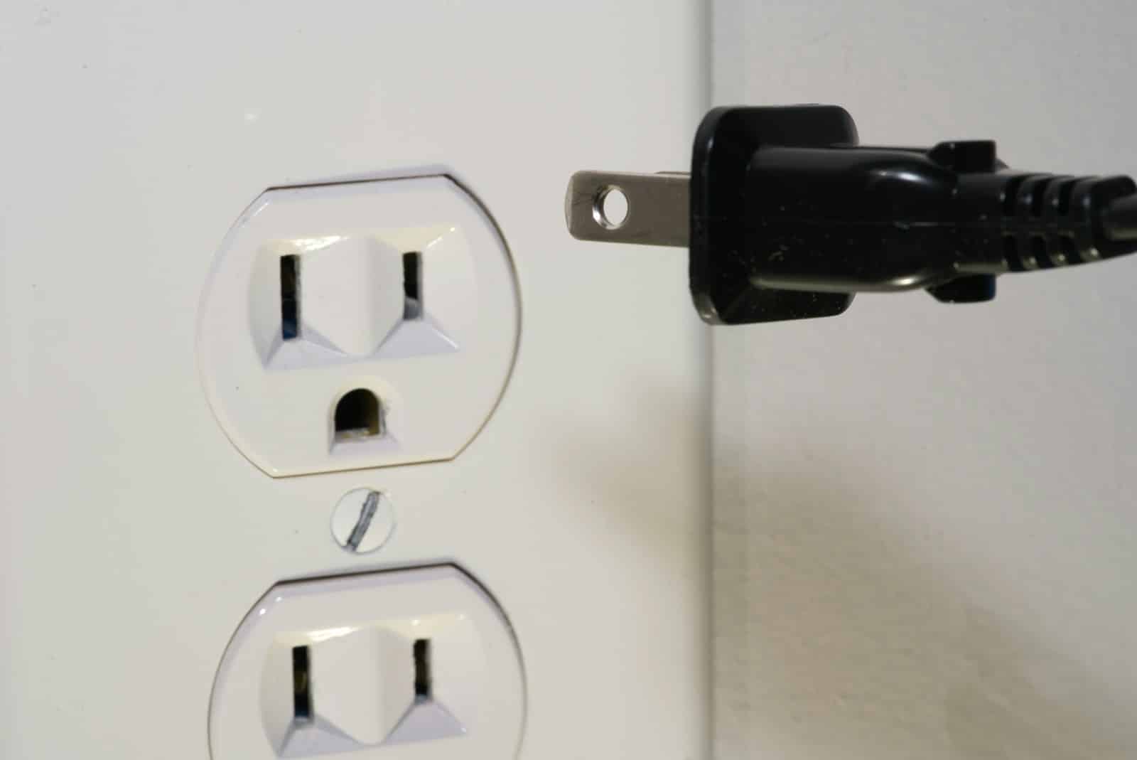 A close-up of a black electrical plug positioned near a white wall outlet, ready to be plugged in. The outlet has two sockets and is mounted on a white wall. black male plug in front of electric socket
