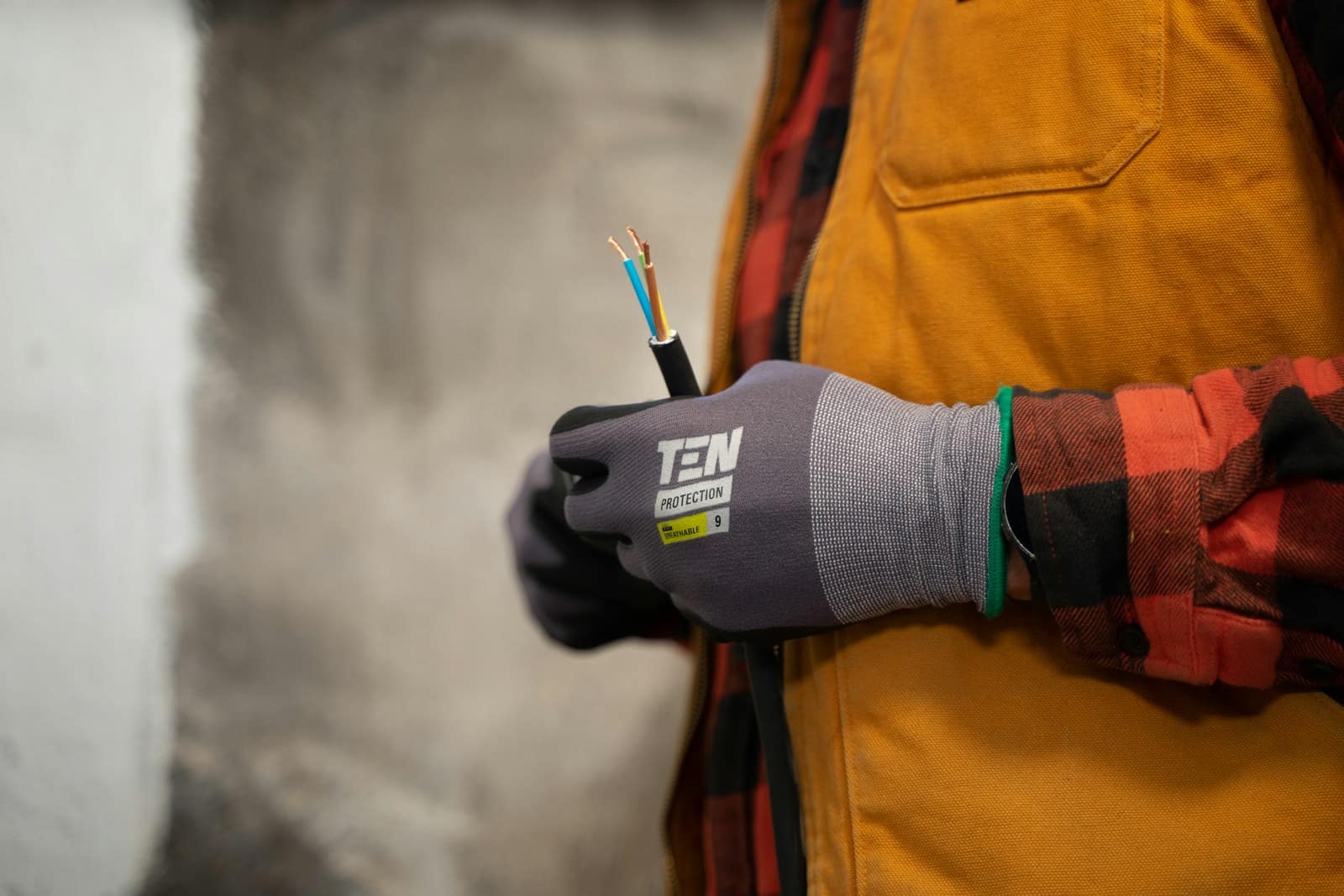 A person wearing gray work gloves and an orange safety vest holds a cut electrical wire with exposed blue and brown wires. The background is blurred. a person holding a pair of wires in their hands